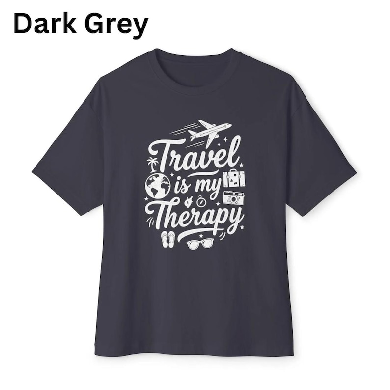 May include: Dark grey t-shirt with the text "Travel is my Therapy" in white. The design includes a plane, globe, camera, and other travel-related icons. The shirt is a crew neck style.