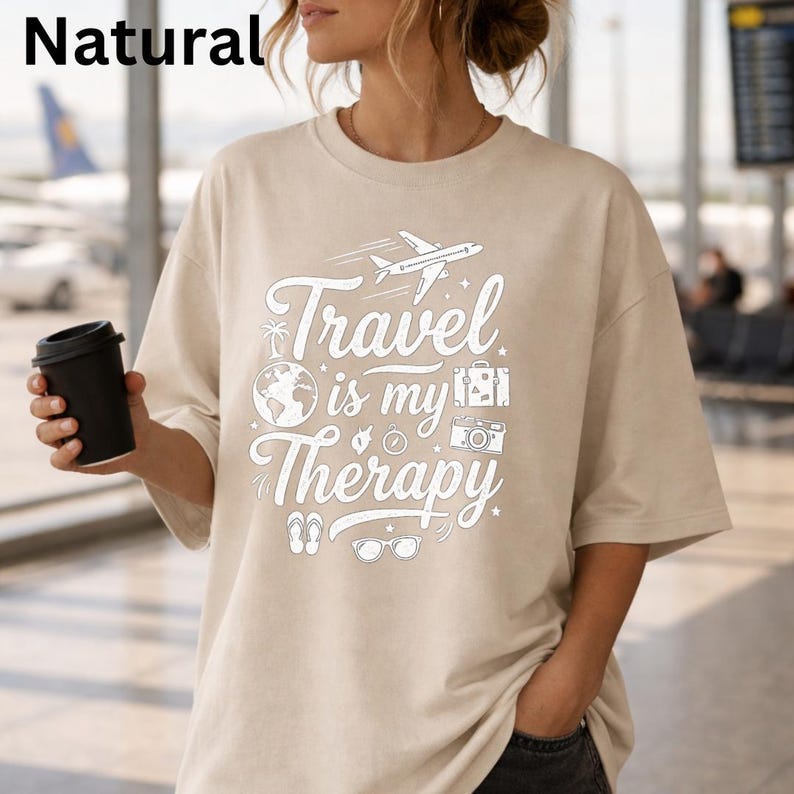 May include: Beige t-shirt with the text "Travel is my Therapy" in white, featuring travel-themed illustrations. The shirt has short sleeves and a relaxed fit. A black coffee cup is held in the left hand.