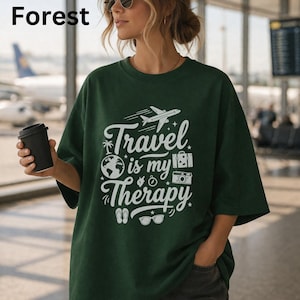 May include: Forest green t-shirt with the text "Travel is my Therapy" in white. The graphic includes a plane, globe, suitcase, camera, sunglasses, and flip-flops. The word "Forest" is in the top left corner.