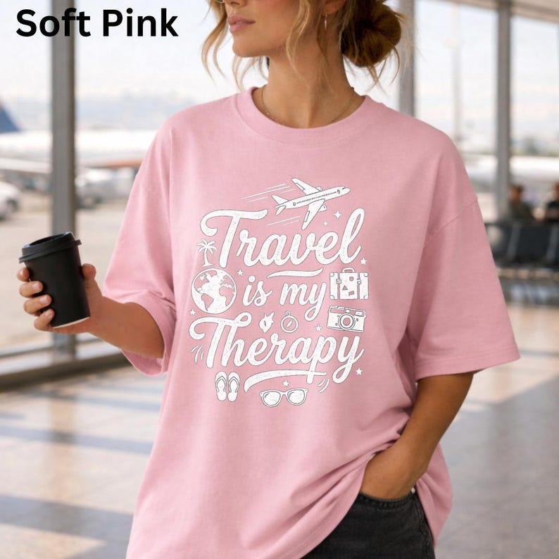 May include: Soft pink t-shirt with the text "Travel is my Therapy" in white. The design includes a plane, globe, suitcase, camera, sunglasses, and flip-flops. The shirt is a soft pink color.