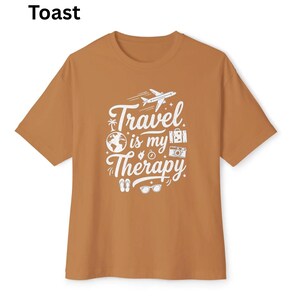 May include: A short-sleeved, toast-colored t-shirt with the words "Travel is my Therapy" in white script and graphics. The design includes an airplane, globe, camera, and other travel-related icons. The word "Toast" is printed in the upper left corner.