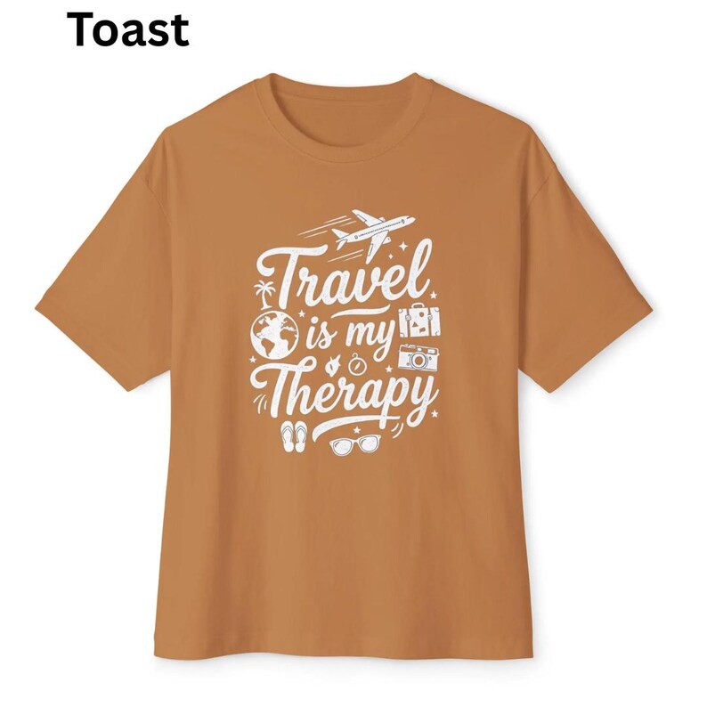 May include: A short-sleeved, toast-colored t-shirt with the words "Travel is my Therapy" in white script and graphics. The design includes an airplane, globe, camera, and other travel-related icons. The word "Toast" is printed in the upper left corner.