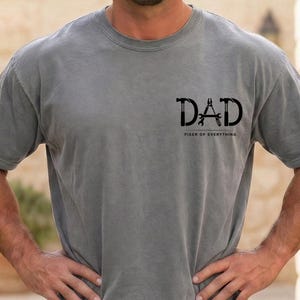 Könnte beinhalten: Graues T-Shirt mit dem schwarzen Wort "DAD", das Hammer- und Schraubenschlüssel-Grafiken enthält. Unter dem Wort "DAD" steht der Text "FIXER OF EVERYTHING".
