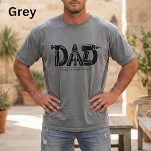 Könnte beinhalten: Graues T-Shirt mit dem Wort "DAD" in schwarzer Schrift, das Werkzeuggrafiken enthält. Unter dem Wort "DAD" steht der Text "FIXER OF EVERYTHING". Das Shirt hat eine einfarbige graue Farbe.