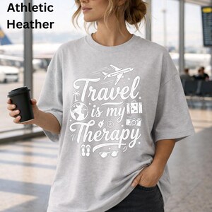 May include: Athletic heather gray t-shirt with the text "Travel is my Therapy" in white. The design includes a plane, globe, suitcase, camera, sunglasses, and flip-flops. The shirt is a relaxed fit.