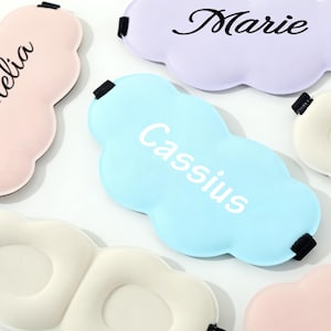 May include: A collection of cloud-shaped sleep masks in various pastel colors, including pink, blue, and lavender. Each mask features a name in a contrasting font: Amelia, Cassius, and Marie. The masks have black elastic straps.