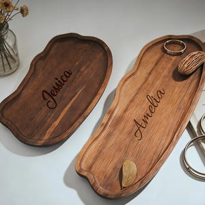Pode incluir: Duas bandejas de madeira para servir com bordas orgânicas. Uma é marrom escuro e gravada com "Jessica", a outra é mais clara com "Amelia". Um pequeno vaso de vidro com flores secas e anéis dourados estão por perto.