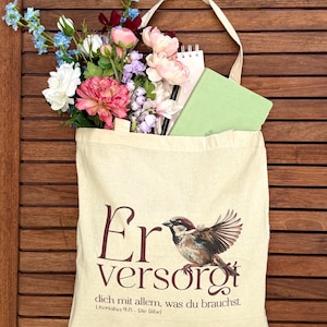 Könnte beinhalten: Eine beigefarbene Stofftasche mit einer Illustration eines Spatzen und dem deutschen Text "Er versorgt dich mit allem, was du brauchst. 1.Korinther 9,8 – Die Bibel." Die Tasche ist mit einem Strauß aus blauen, rosa und weißen Blumen sowie einem hellgrünen Notizbuch gefüllt.