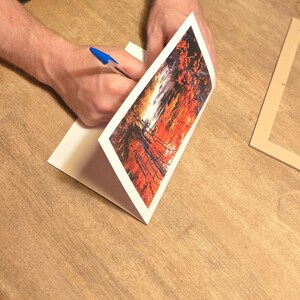 May include: A person is writing on a white card with a blue pen. The card features a vibrant abstract art print in shades of red, orange, and black. A wooden frame is on the table.