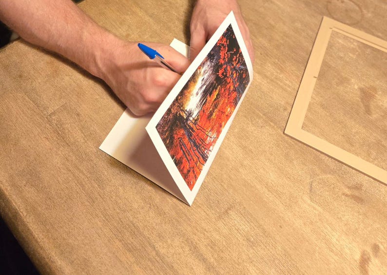 May include: A person is writing on a white card with a blue pen. The card features a vibrant abstract art print in shades of red, orange, and black. A wooden frame is on the table.