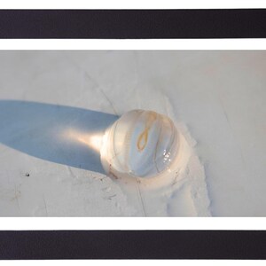 May include: A framed photograph featuring a close-up of a translucent, spherical object with a swirling, marbled pattern in shades of white and gold. The object casts a long shadow on a light-colored surface.