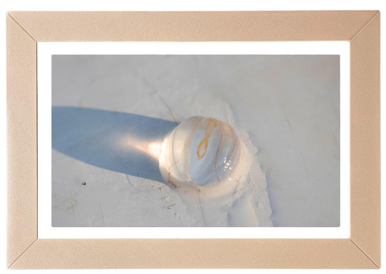 May include: A framed photograph featuring a close-up of a translucent, spherical object with a light-colored, textured surface. The object casts a long shadow on a light-colored surface. The frame is a light beige color.