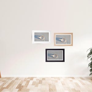 May include: Three framed prints of a glass object on a sandy surface, each with a different frame color: white, tan, and black. The prints are mounted on a white wall, with a wooden floor and a floor lamp with a white shade.