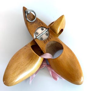 May include: A pair of vintage wooden shoe forms, light brown in color, with a metal hinge and ring. The forms are tied together with a pink ribbon. The shoe forms have a rounded toe and a circular opening on the side.