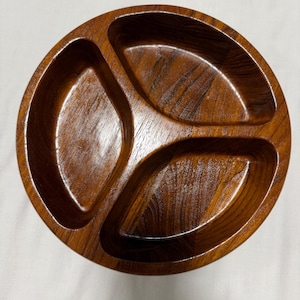 May include: A round, three-sectioned wooden serving dish with a rich, dark brown finish. The dish has a glossy sheen, highlighting the natural wood grain. The sections are oval-shaped, perfect for serving snacks or appetizers.