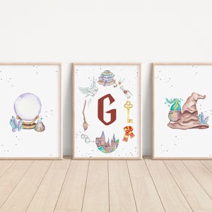 Może przedstawiać: Trzy oprawione wydruki akwarelowe o tematyce magicznej. Wydruki przedstawiają kryształową kulę, dużą literę "G" z magicznymi symbolami oraz tiarę z bulgoczącym eliksirem. Grafika jest na białym tle.