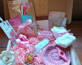 May include: A gift box filled with pink-themed items. Includes a crochet coaster, a book, tea, a face massager, a bookmark, a paper flower, and other small gifts. The box is lined with pink paper shreds.