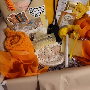 May include: A gift box filled with orange and yellow items, including a bookmark with the text "BOOK NERD", pens, a crocheted item, face masks, and tea bags. The box is lined with brown paper and orange tissue paper.
