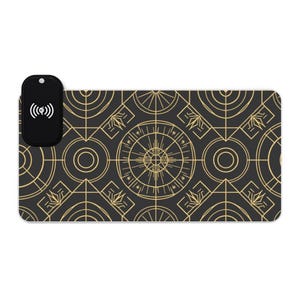 May include: A rectangular desk mat featuring a black background with a repeating gold geometric pattern of circles, lines, and starbursts. A black wireless charging module with a white Qi logo is attached to the top left corner of the mat.