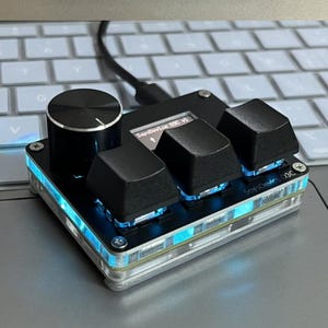 May include: A black and silver electronic device with a large knob, three black keys, and a small screen. The device has a clear, illuminated border with blue lights. The text "Sayodevice OSC V1" is visible on the screen.