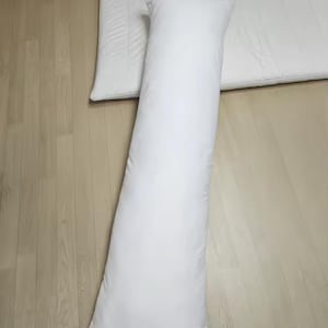 Puede incluir: Una almohada corporal blanca, larga y rectangular, colocada verticalmente junto a un protector de colchón blanco, plano y rectangular. Ambos artículos presentan una superficie de tela blanca lisa y sólida con bordes rectos.