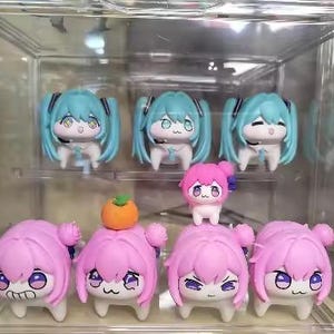 May include: A selection of anime-style figurines displayed in a clear case. The figures have large eyes and stylized hair in pink and teal. Some have small orange fruit accessories. The figures are approximately 5-7 cm tall.