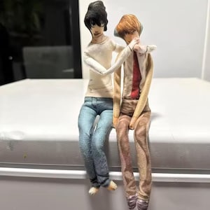 May include: Two handmade fabric dolls are seated together. One has black hair, a white shirt, and blue jeans. The other has brown hair, a white shirt, a red tie, and tan trousers. The dolls appear to be made of fabric.