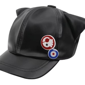 Könnte beinhalten: Schwarze Baseballkappe mit Katzenohren und gebogenem Schirm. Die Kappe hat zwei runde Anstecknadeln: eine mit einem rot-weißen Design und eine weitere mit einem blau-weißen Zielscheibenmuster. Die Kappe ist aus glattem, möglicherweise lederähnlichem Material.