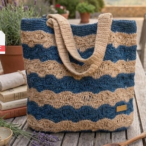 May include: A large, handmade tote bag with a blue and tan wave pattern. The bag has two tan handles and a small rectangular tag. The bag is sitting on a wooden table with other items, including books and a bowl.