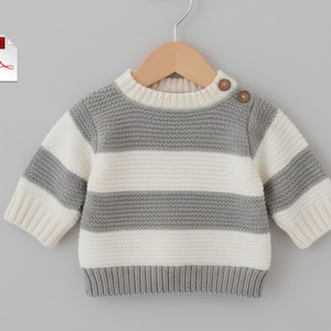 Może przedstawiać: Szary i biały sweter w paski dla dziecka z okrągłym dekoltem i długimi rękawami. Sweter ma dwa brązowe guziki na ramieniu i jest prezentowany na drewnianym wieszaku.