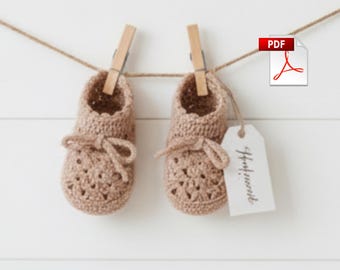 May include: Two light brown, handmade crochet baby booties suspended from a clothesline with wooden pegs. Each bootie features a bow and a tag that reads "Handmade". A PDF icon is in the top right.