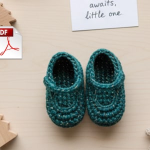 Może przedstawiać: Para turkusowych szydełkowych bucików dla dziecka z zapięciem na guziki, obok drewnianych zabawek i małego misia. Na kartce napis "awaits, little one". Ikona PDF znajduje się w lewym górnym rogu. Buty mają około 10 cm długości.