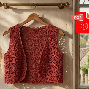 May include: A rust-colored sleeveless crochet vest featuring an intricate openwork pattern throughout the garment. The vest has a rounded neckline and a scalloped edge along the front opening, hanging on a wooden hanger against a wall.