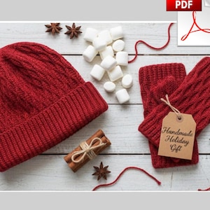 Op de afbeelding: Een rode gebreide muts en bijpassende vingerloze handschoenen met een kabelpatroon. De handschoenen hebben een label met de tekst "Handmade Holiday Gift". De afbeelding bevat ook marshmallows, kaneelstokjes en steranijs.