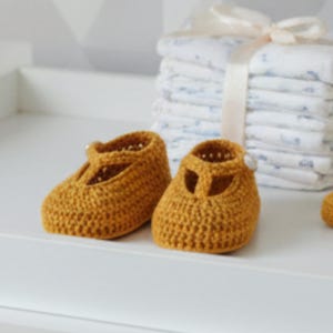Könnte beinhalten: Ein Paar senfgelbe gehäkelte Babyschuhe mit T-Steg-Design. Die Schuhe sind handgefertigt und liegen auf einer weißen Oberfläche. Ein Stapel weißer und blau gemusterter Babywindeln befindet sich im Hintergrund.