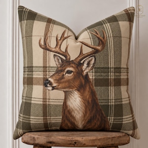 May include: A square throw pillow featuring a detailed illustration of a brown deer with large antlers centered on a beige and olive green plaid fabric pattern. The fabric has a visible herringbone texture throughout the design of the pillow.