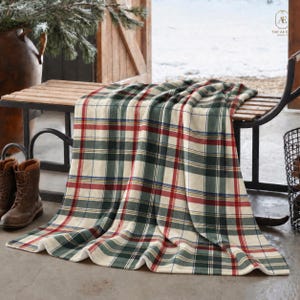 Può includere: Una morbida coperta con un classico motivo a quadri nei toni del verde bosco, crema e rosso scuro con sottili linee di accento blu navy. Il tessuto è drappeggiato su una panca in legno, mostrando una trama tessuta e un bordo rifinito.