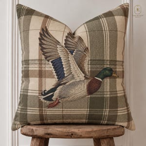 May include: A square throw pillow featuring a woven plaid pattern in shades of olive green, beige, and brown. Centered on the front is a detailed, embroidered illustration of a mallard duck in flight with its wings spread, showing brown, blue, and green plumage.