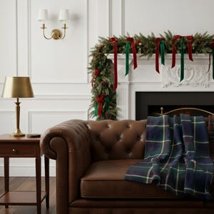 May include: A cozy living room scene featuring a brown leather Chesterfield sofa with a blue, green, and white plaid blanket. A gold lamp sits on a wooden side table. A fireplace is decorated with a garland and red and green ribbons.