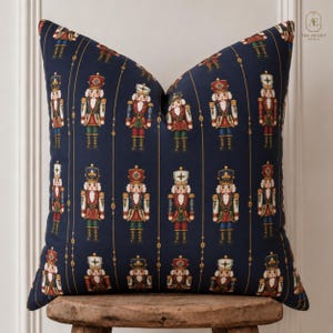 May include: A square throw pillow with a dark navy blue background featuring a repeating pattern of colorful nutcracker figures arranged in vertical columns, separated by thin gold lines. The nutcrackers wear red, green, and blue uniforms.