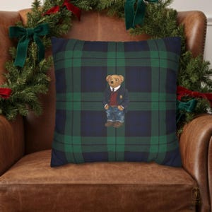 Puede incluir: Cojín decorativo cuadrado con un estampado de cuadros azul marino y verde. El cojín presenta un oso de dibujos animados con blazer, vaqueros y zapatos marrones. El cojín está sobre un sillón de cuero marrón con una corona navideña de fondo.
