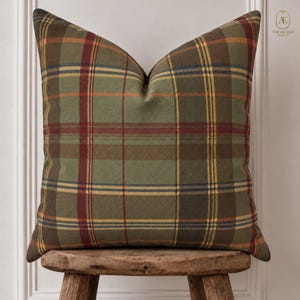 May include: A square scatter cushion featuring a woven plaid pattern in shades of olive green, deep red, mustard yellow, and navy blue. The fabric has a visible herringbone texture throughout, and the cushion is displayed on a rustic wooden stool.