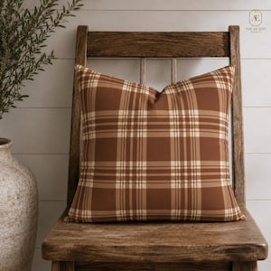 Peut inclure: Un coussin carré avec un motif à carreaux dans des tons de brun, de beige et de crème. Le coussin présente une texture de tissu lisse et uniforme et est posé sur une chaise en bois. Le motif se compose de lignes horizontales et verticales.