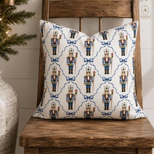 May include: A square throw pillow featuring a repeating pattern of nutcracker figures in blue and red uniforms, each holding a peppermint staff. The nutcrackers are framed by a lattice design of light blue bows on a white background.