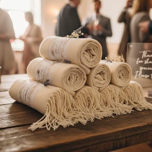 Puede incluir: Una pila de chales color crema enrollados con bordes con flecos, decorados con pequeños detalles florales. Los chales están colocados sobre una mesa de madera rústica, con un letrero transparente que dice "Thank you for sharing our special day. Please take one."
