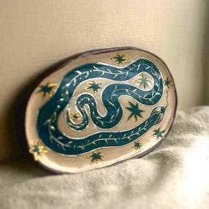 Puede incluir: Bandeja ovalada de cerámica con un diseño de serpiente turquesa y detalles de estrellas doradas. La serpiente serpentea sobre la superficie beige, rodeada de estrellas doradas y delicados detalles de vid. La bandeja tiene un borde marrón oscuro.