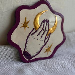 May include: A decorative ceramic plaque with an irregular shape, featuring a cream-colored center and a deep purple border. The design displays a black-lined illustration of a hand cradling a gold crescent moon, accented by two gold stars.