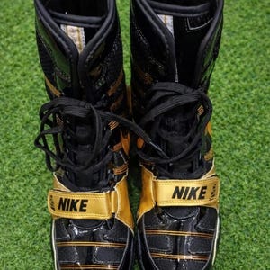 May include: Pair of black and gold Nike wrestling boots. The boots have a high-top design with a gold strap across the top that says "NIKE". The boots have black laces and gold accents.