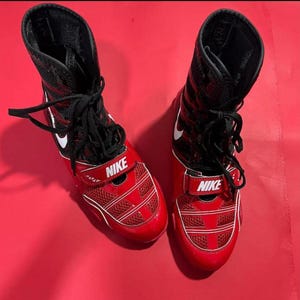 May include: Pair of red and black Nike wrestling shoes. The shoes have a high-top design with black laces and a white Nike swoosh. The front of the shoe features a red strap with the Nike logo.