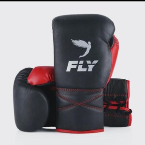 Può includere: Guantoni da boxe neri e rossi con la scritta "FLY" in lettere bianche e una grafica ad ala bianca. I guantoni hanno cuciture e dettagli rossi. I guantoni sono progettati per la boxe o altri sport da combattimento.